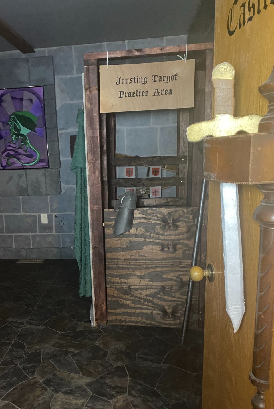 Castle-themed room with a stone-look wall, a dragon painting, a jousting practice booth and a sword by the door