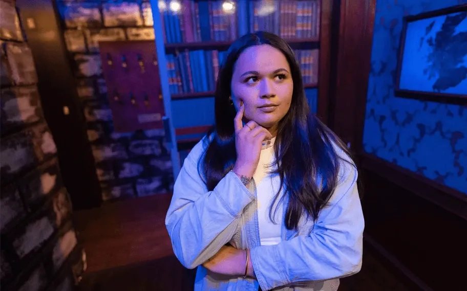 A player deep in thought in the Mansion Murder room, with bookshelves and blue patterned wallpaper behind her