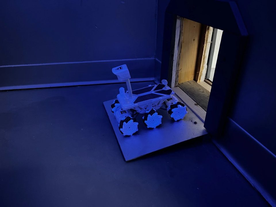 A toy Mars rover on a platform next to a small hatch in a blue-lit room