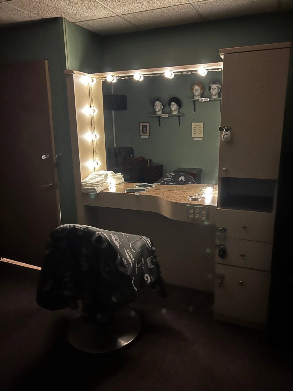 Barbershop set with a lit vanity mirror, a draped barber chair and wig heads on the shelves