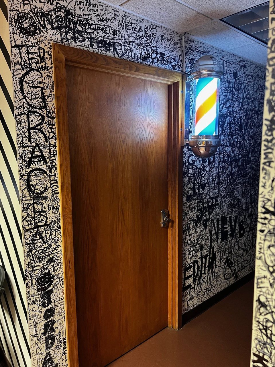 Entrance to the barbershop room: a wooden door and a lit barber pole on a wall covered in handwritten names