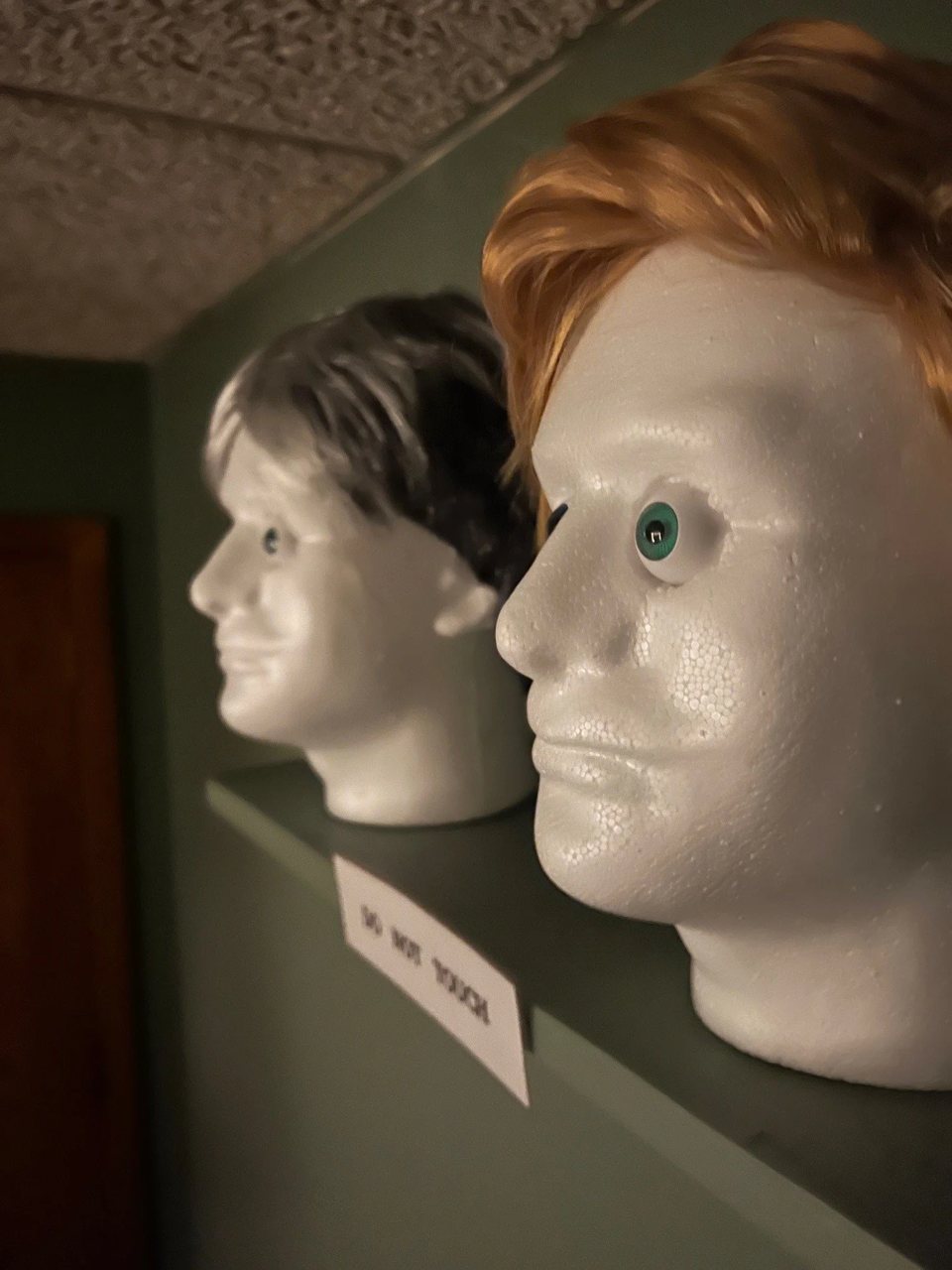 Two foam mannequin heads with wigs and a Do Not Touch sign on a shelf