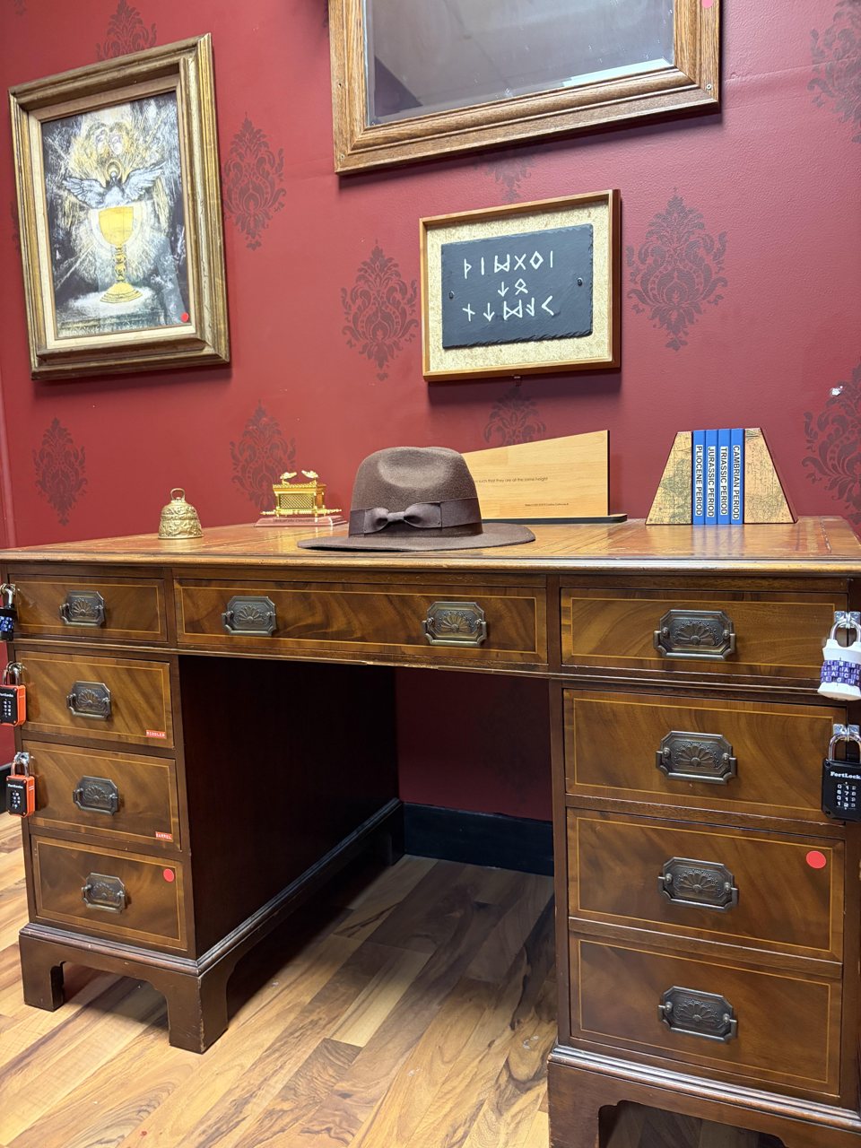 Antique desk with a fedora, padlocked drawers and framed clues on the red wall above