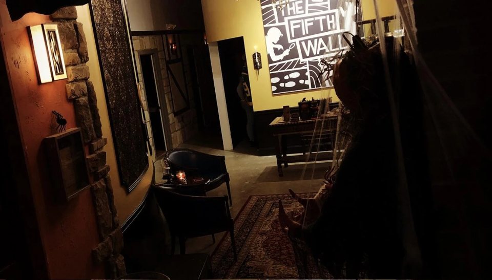 Félhomályos előtér karosszékekkel, perzsaszőnyeggel, kőfallal és nagy Fifth Wall falképpel, előtérben egy fátyol mögötti jelmezes alakkal