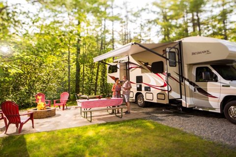 An RV on a wooded campsite with lawn chairs