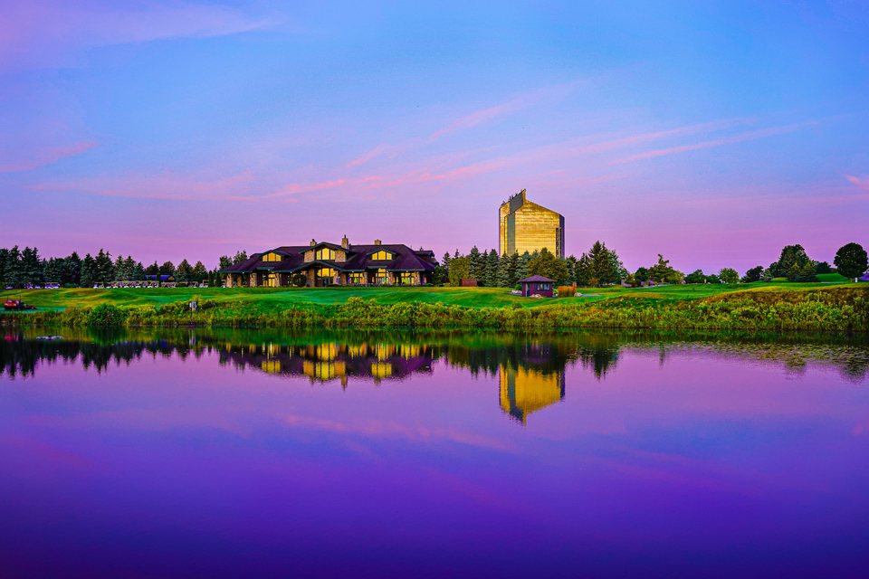 The resort's golden hotel tower and a low timber building reflected in a pond at purple dusk