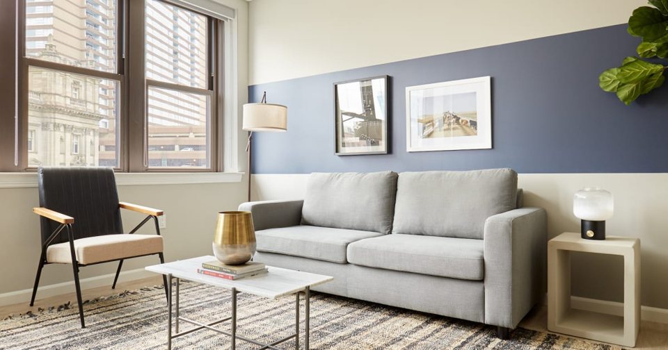 An apartment living room with a gray sofa and windows facing downtown buildings