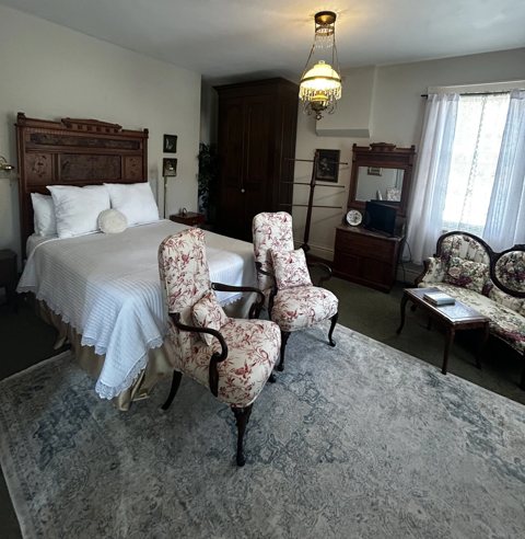 A spacious guest room with a carved antique bed, floral armchairs and a blue patterned rug
