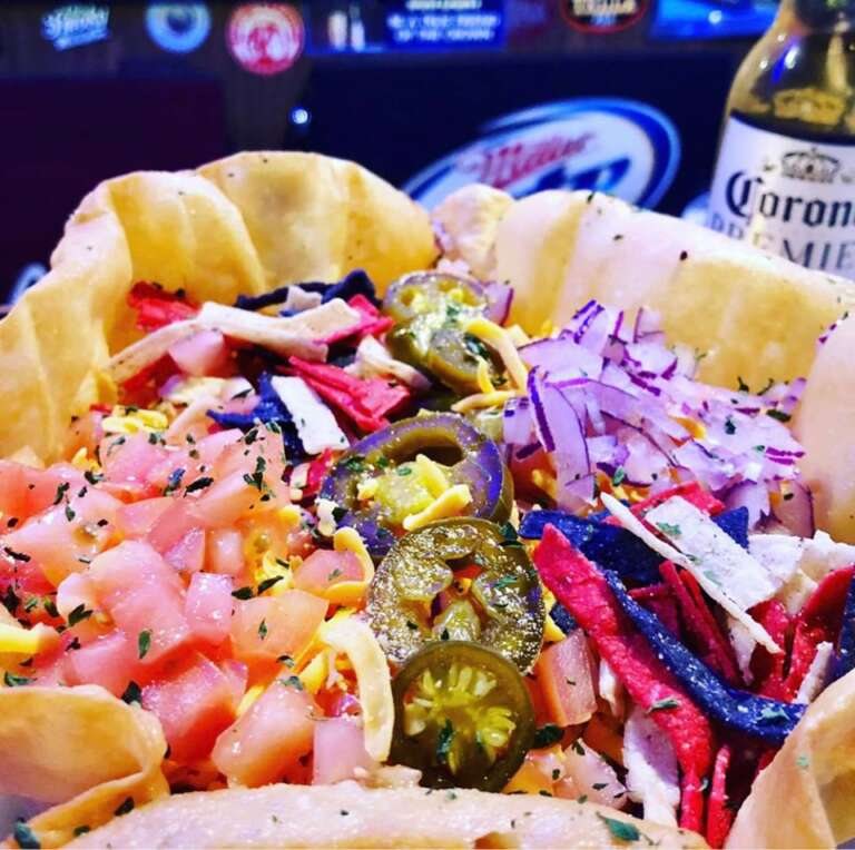 Tacosaláta tortillatálban paradicsommal, jalapeñóval, lilahagymával és tortillacsíkokkal, mögötte sörreklámok és egy sörösüveg