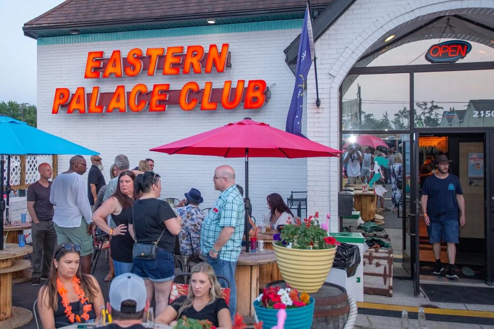 Fehér épület piros Eastern Palace Club felirattal, előtte napernyős terasz beszélgető vendégekkel