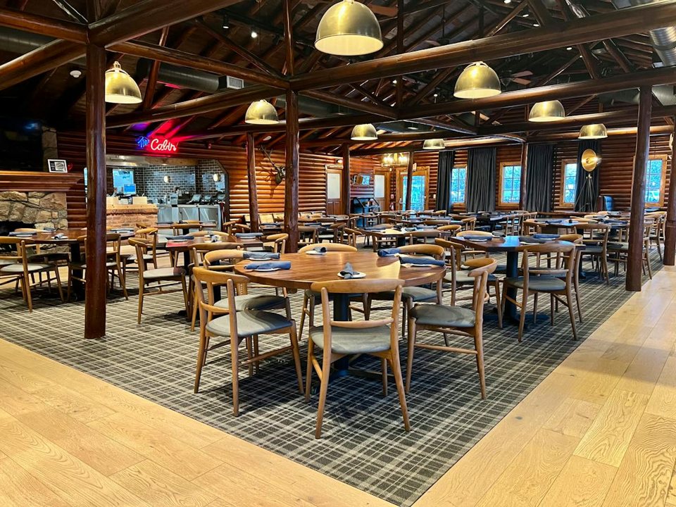A log-walled dining room with a beamed ceiling, round tables, a plaid rug and pendant lamps