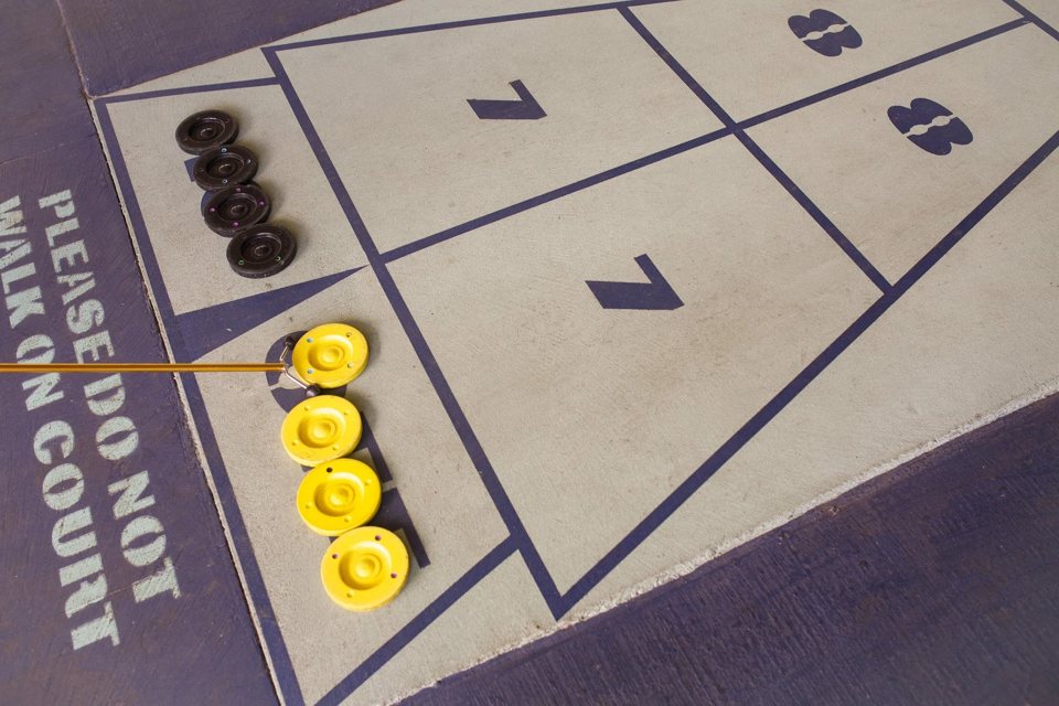 Shuffleboardpálya közelről, számozott mezőkkel és sárga, fekete korongokkal