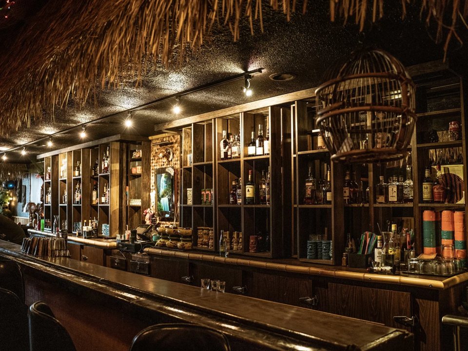 A long dark wooden bar with liquor shelves behind it under a thatched overhang