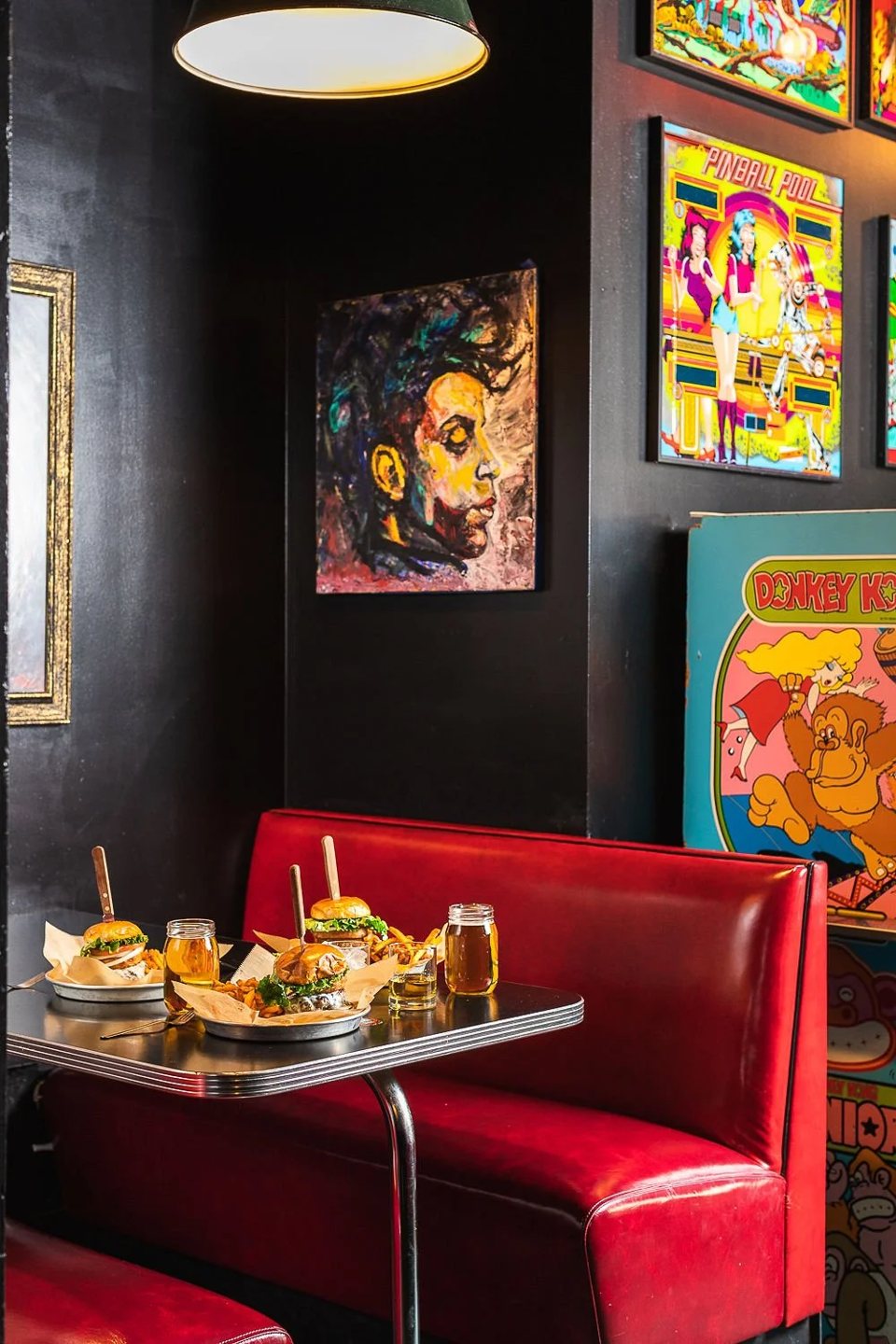 A red vinyl booth with burgers and beer on the table and paintings on the wall