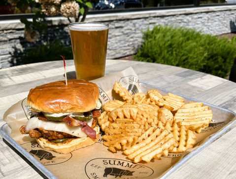 A bacon burger with waffle fries on Summit-branded paper, with a glass of beer behind it on the patio