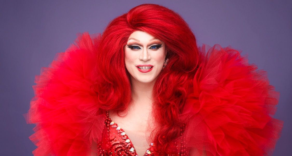 Vörös parókás drag előadó dús, vörös tüllgallérral, lila háttér előtt