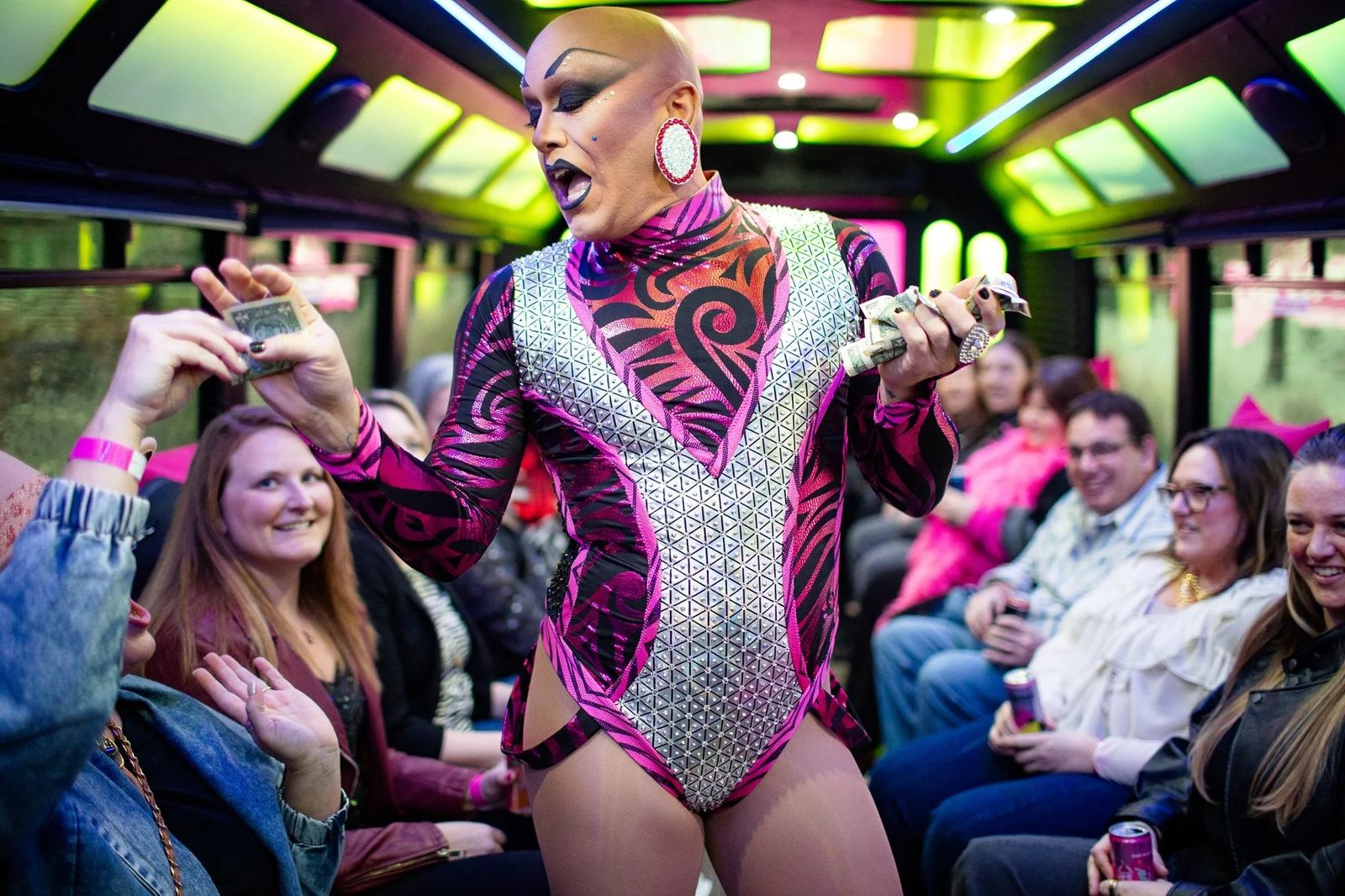 Drag performer in a sparkly pink bodysuit collects tips from cheering bus riders
