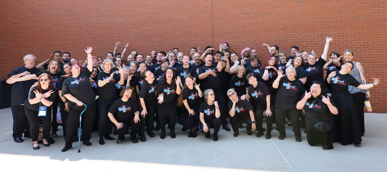 Out Loud Chorus singers in black T-shirts pose cheerfully in front of a brick wall