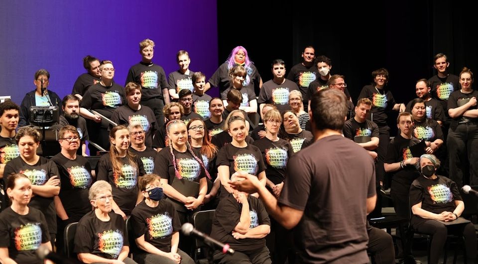 Chorus members in rainbow-print T-shirts on stage, facing their director