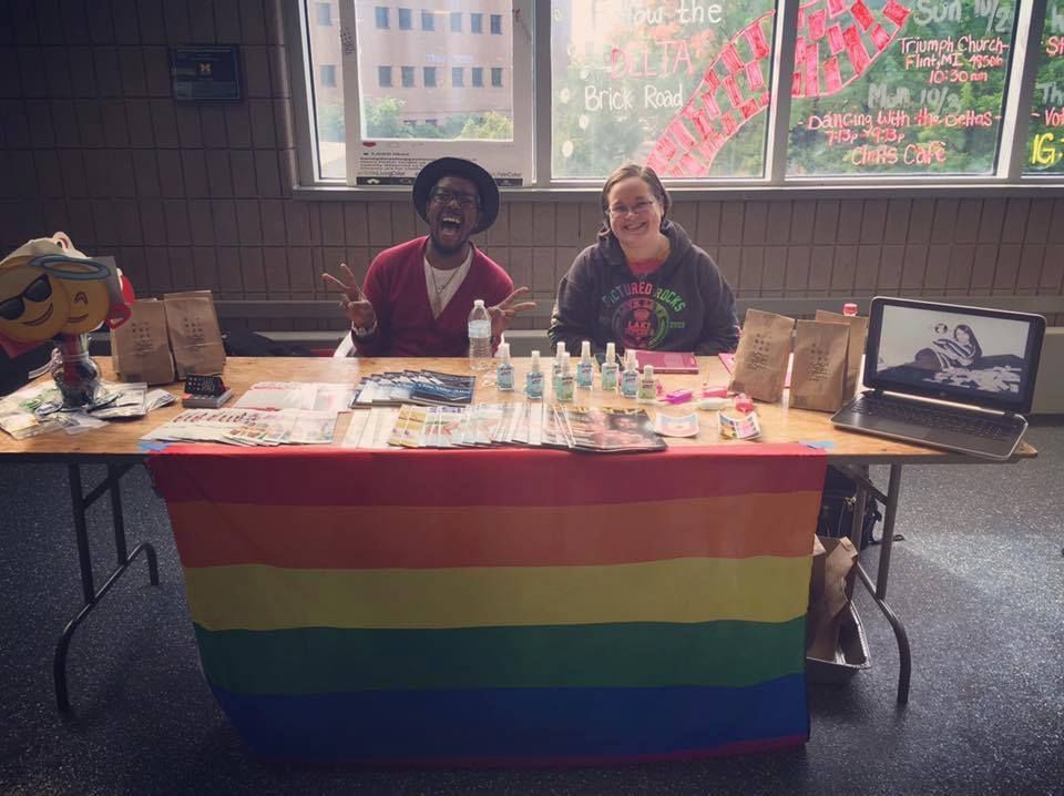 Two smiling volunteers at an outreach table draped with a rainbow flag