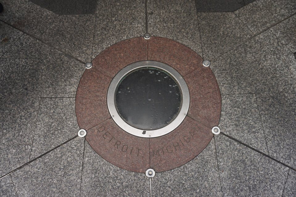 The Point of Origin medallion set into the stone paving of Campus Martius Park: a brown granite ring, a glass porthole in the middle, the words Point of Origin, Detroit, Michigan around it