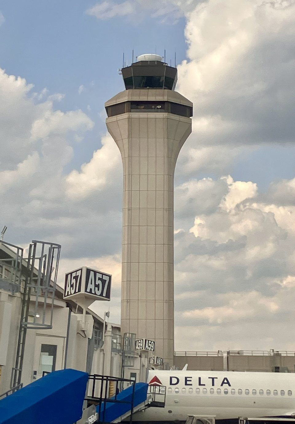 A Detroit Metro Airport légiforgalmi irányítótornya gomolyfelhős ég előtt; alatta egy Delta-gép áll az A57-es kapunál.