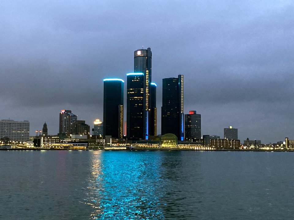 Kék fényben ragyogó tornyok és tükörképük a vízen: a Renaissance Center Windsorból, 2023. december 31-én alkonyatkor
