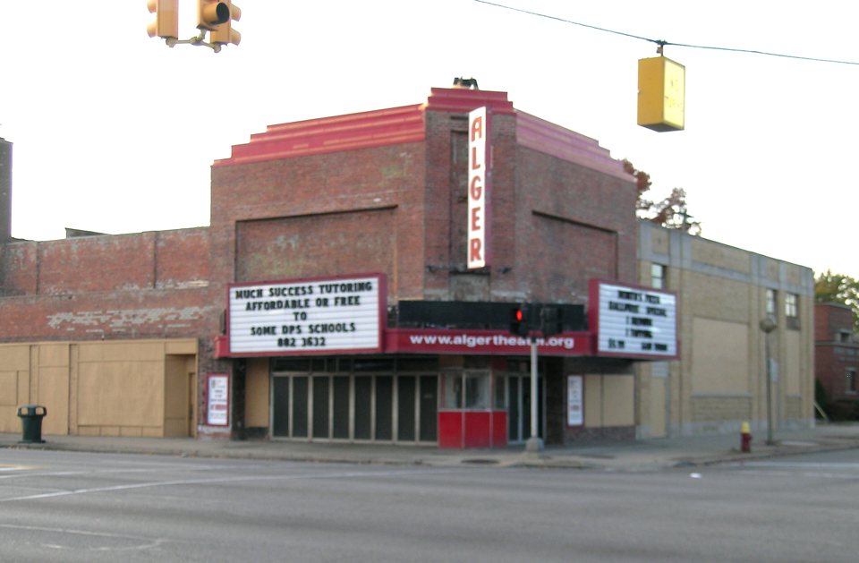The Alger Theater's red-brick facade with its vertical ALGER sign, two marquee boards and boarded-up storefronts at the intersection