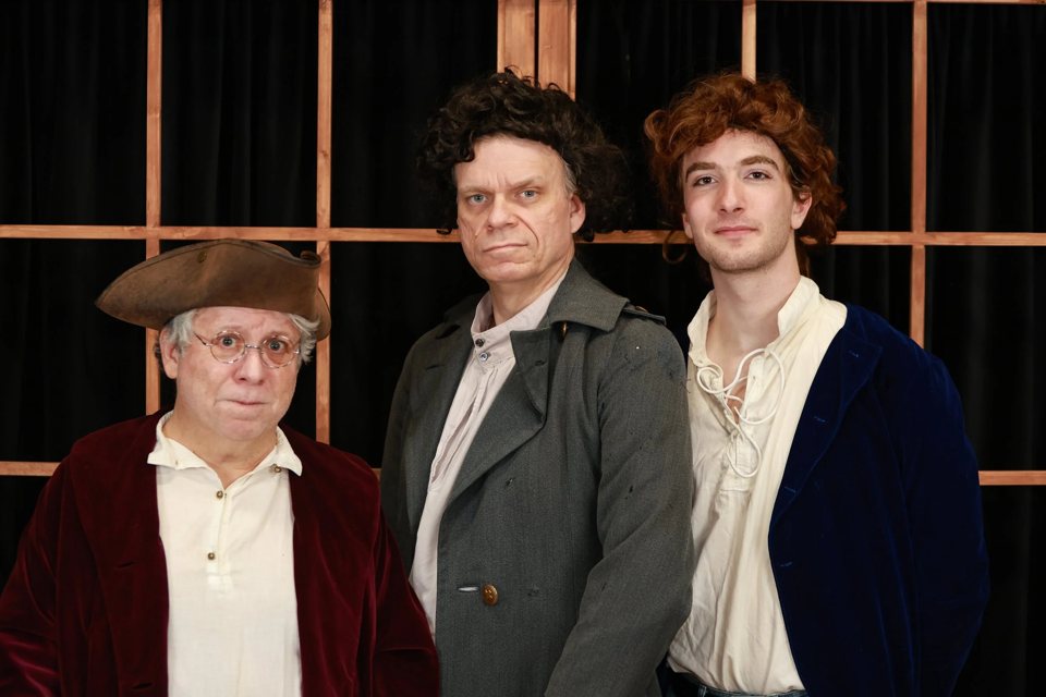 Three actors in period costume, with a tricorn hat, velvet coats and wigs, in front of a wooden-frame set