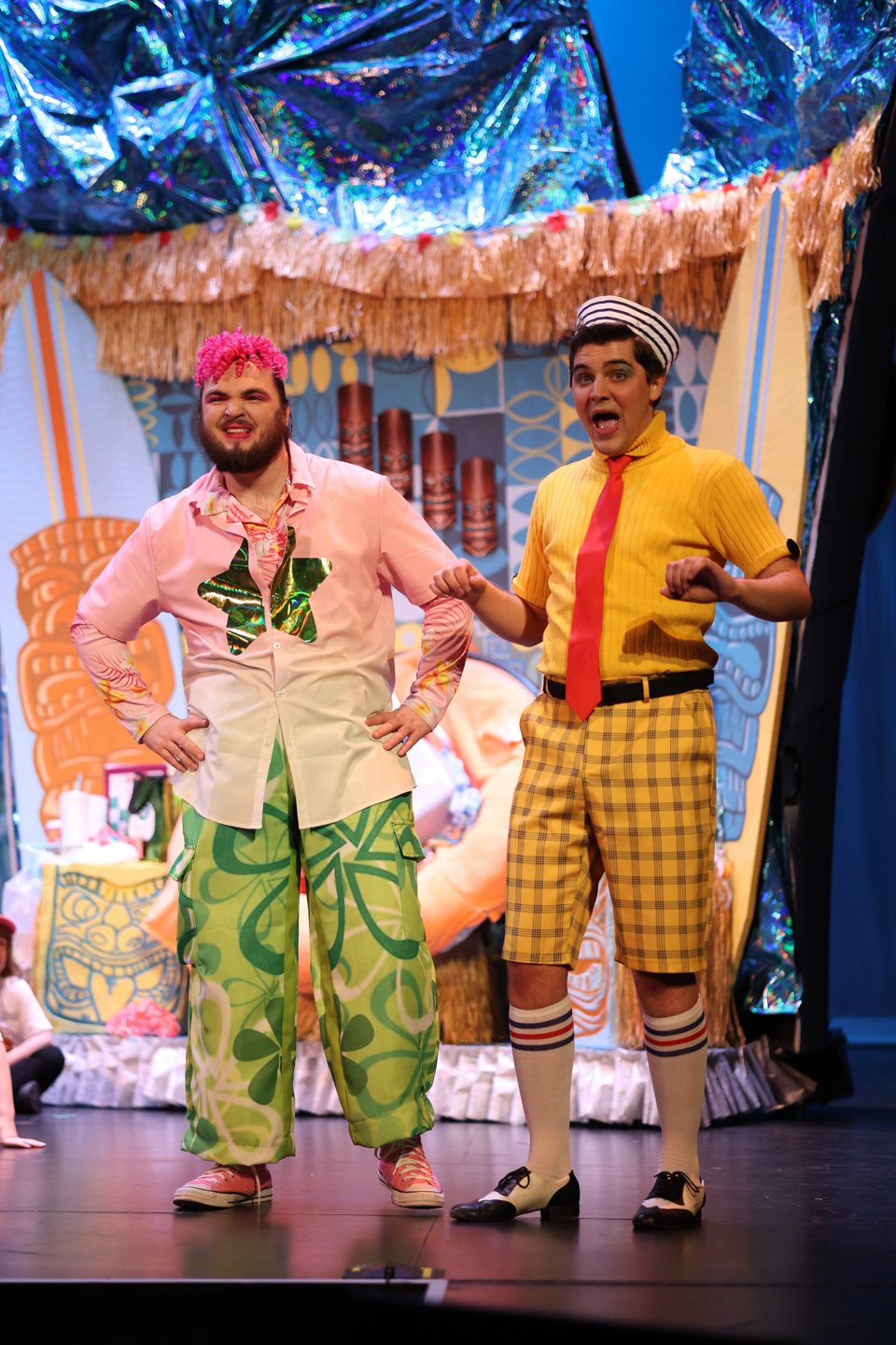 Two actors in loud beach costumes, one in a pink curly wig and green patterned trousers, the other in a yellow shirt, red tie and plaid shorts, in front of a tiki-themed set