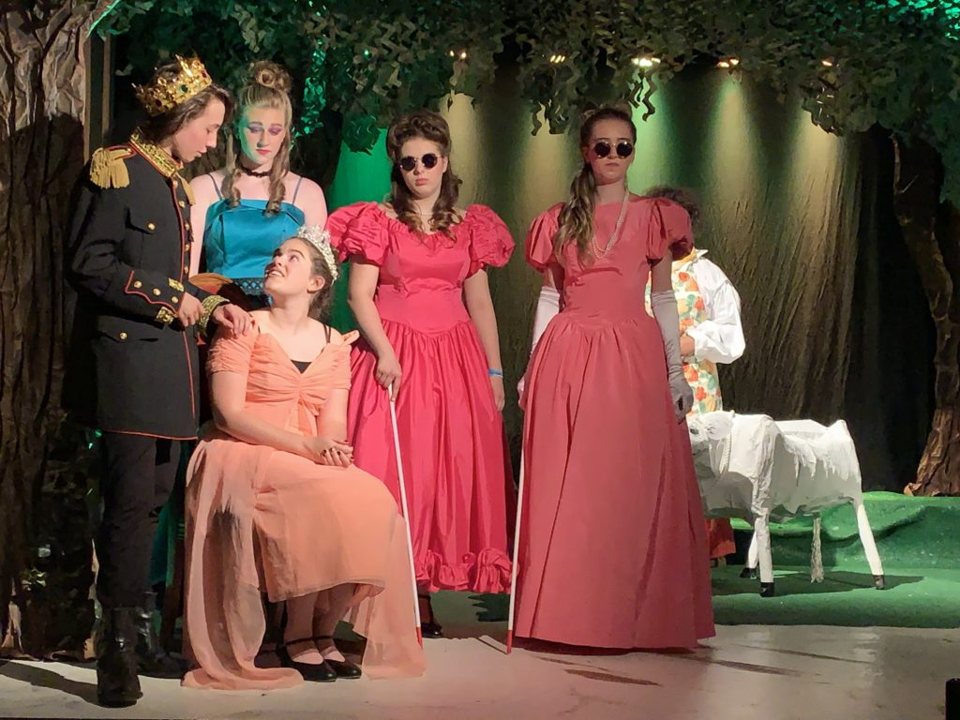 Fairy-tale characters on a forest set: a prince in a crown, a girl in a peach gown, two sisters in pink gowns with dark glasses and white canes, and a white cow prop