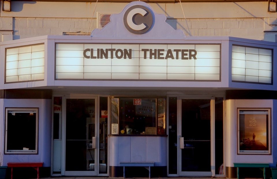 The Clinton Theater's glowing marquee with its CLINTON THEATER lettering and C crest above the ticket booth and glass doors in warm, low sunlight