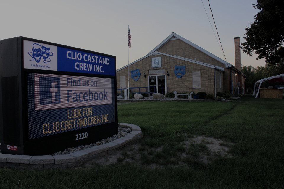 Clio Cast and Crew's yellow-brick theatre with blue theatre masks on the wall and the company's lit sign in front at dusk