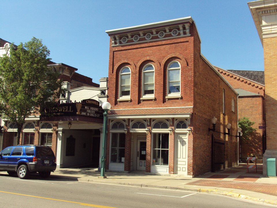 A Croswell Opera House utcai frontja nappal: a régi fekete márkiz a Will Rogers Follies műsorával, mellette a vörös téglás szomszéd ház