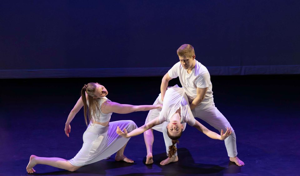 Three dancers in white perform a contemporary piece on a violet-lit stage, one of them arching backwards