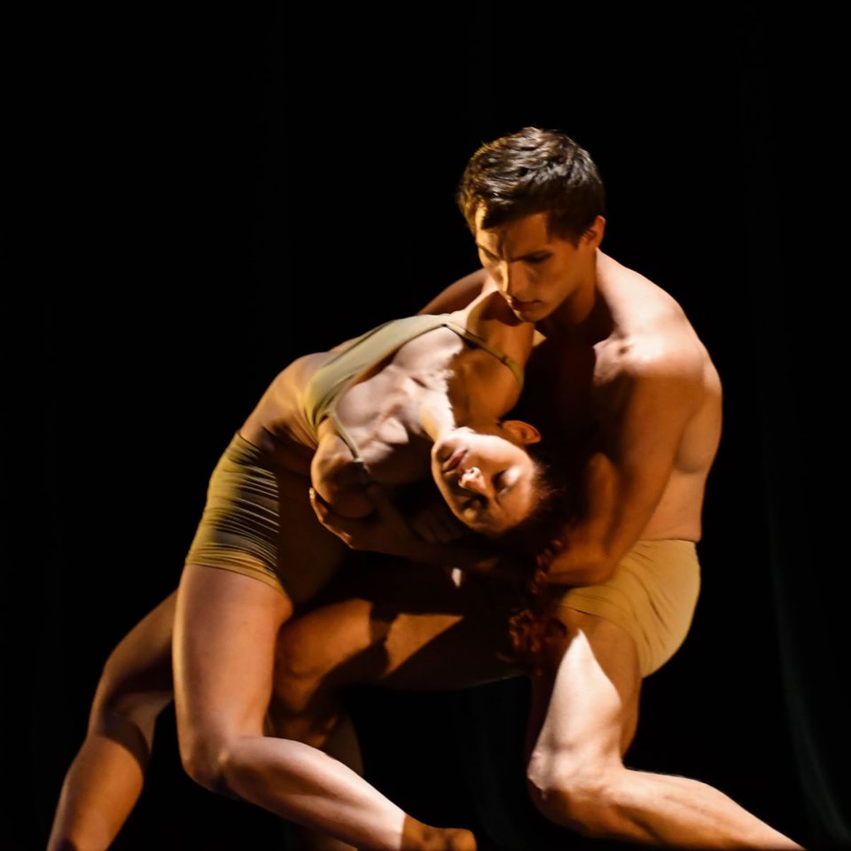 Two dancers in nude-toned costumes, the man holding the woman as she bends back against a dark background