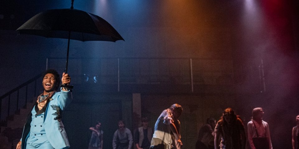 An actor in a light blue suit sings under a black umbrella on a hazy, colorfully lit stage with the ensemble behind him