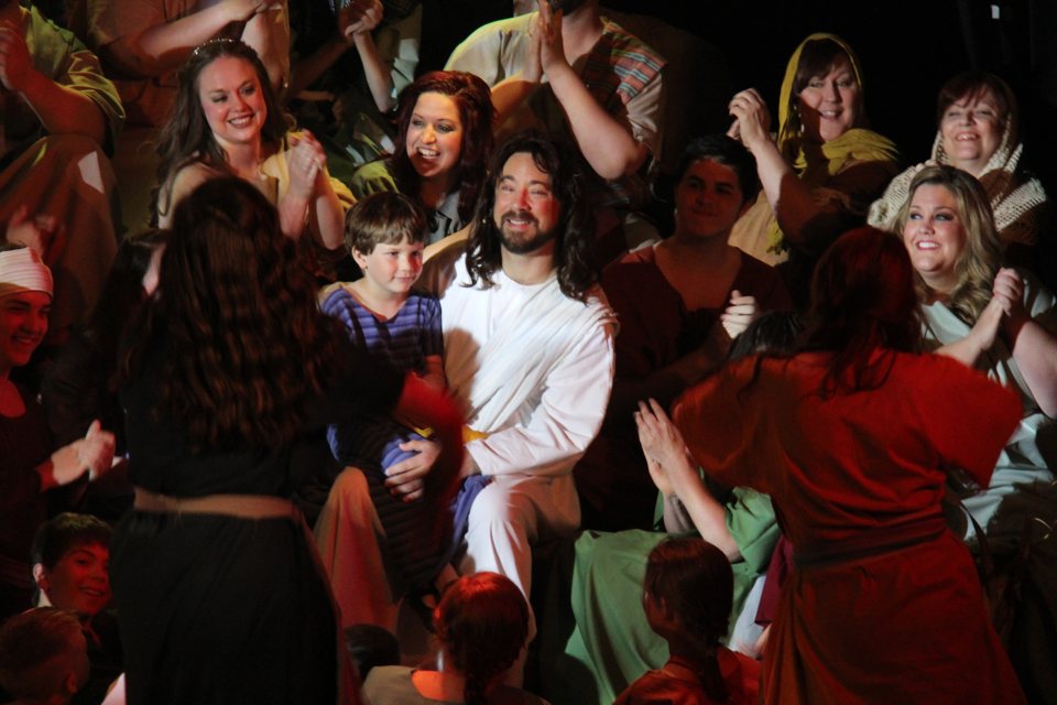 A white-robed Jesus figure holds a young boy on his lap while cast members in biblical costumes clap around them on stage