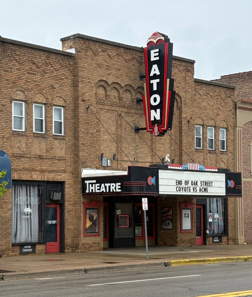 The brick facade of the Eaton Theatre with its red and black vertical EATON sign and a marquee listing films, seen from the street