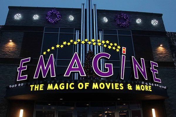 The purple EMAGINE letters, arc of stars and yellow 'The Magic of Movies & More' lettering at dusk, with holiday decorations on the facade
