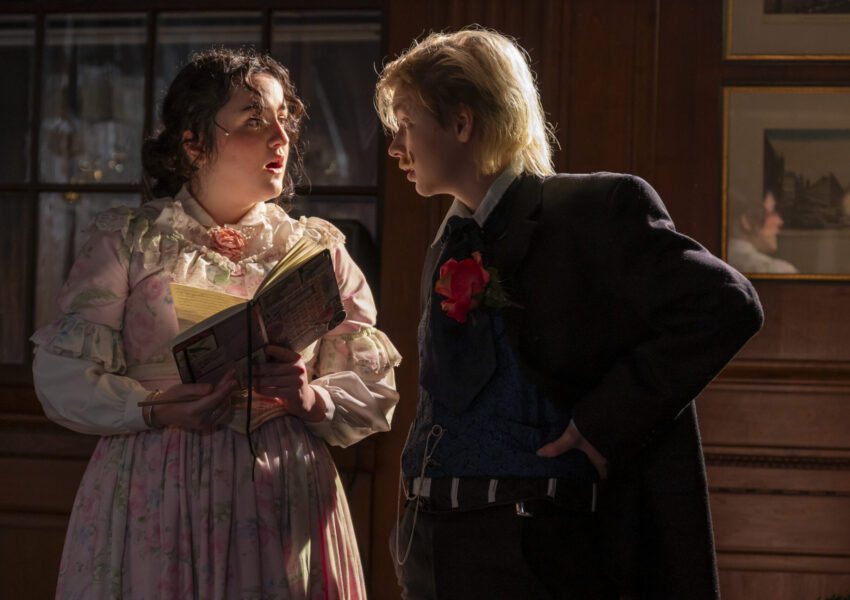 An actress in Victorian costume holding a book talks with a young man in a dark coat and red rose boutonniere on a wood-paneled set