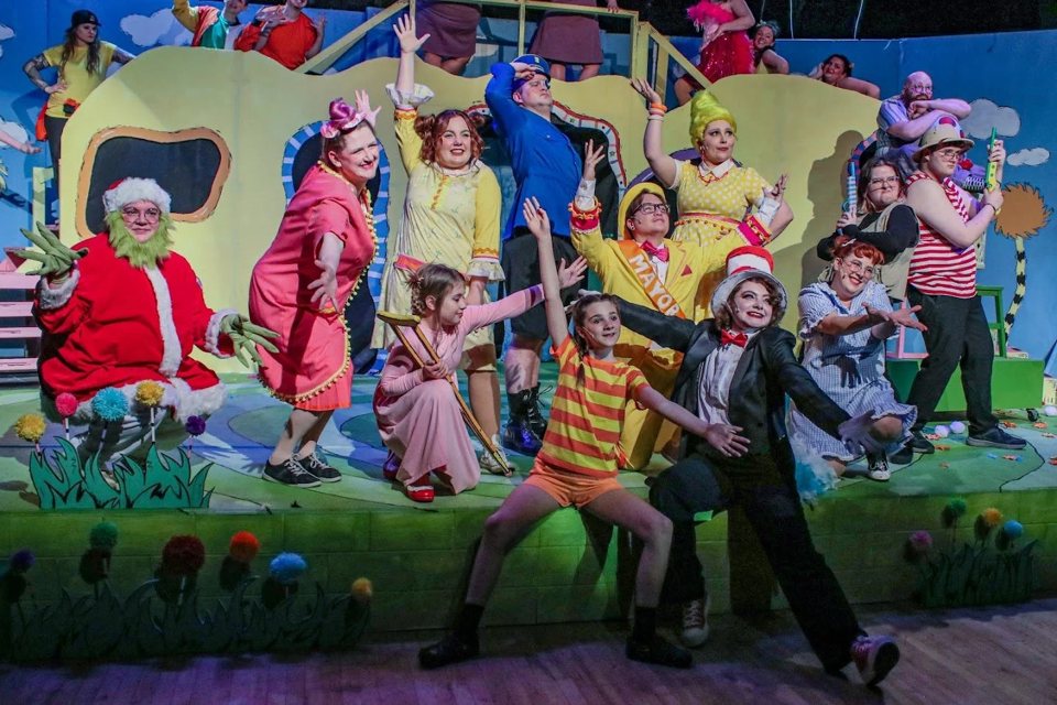 A costumed cast strikes a pose on a colorful storybook set, led by a performer in a red-and-white striped top hat and a girl in an orange striped shirt with arms spread wide
