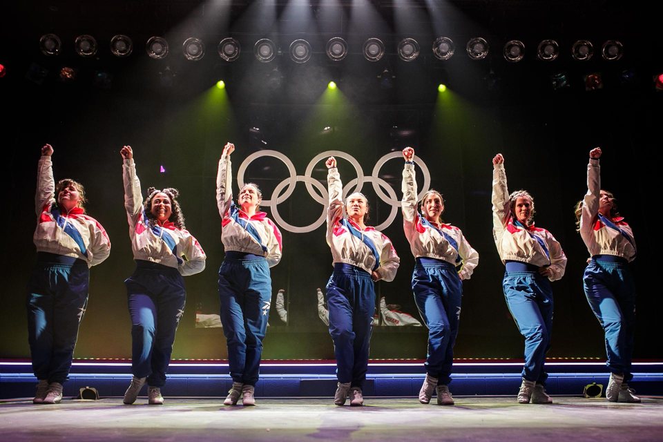 Hét színésznő egyforma kék-fehér-piros melegítőben, felemelt ököllel áll a színpadon az olimpiai karikák előtt, fölöttük reflektorsor