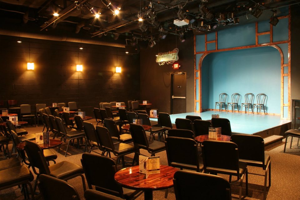 The Go Comedy! house with round tables and black chairs facing the blue stage in its wooden arch, with four chairs set out