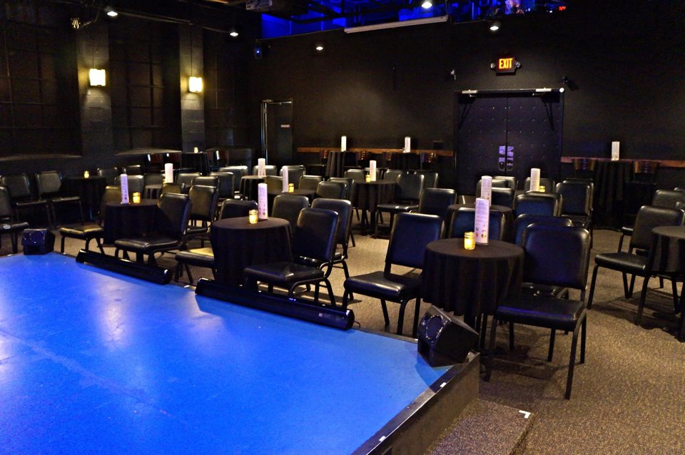 The edge of a blue stage with rows of chairs and candlelit, black-draped tables in the dark-walled room