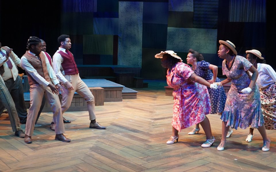 Men in vests face women in floral dresses and hats on a stage with a herringbone wood floor