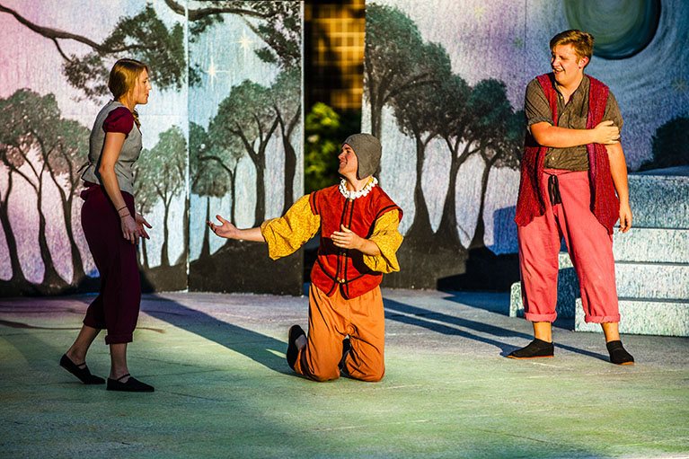 Three costumed actors on stage in front of a painted forest backdrop, the one in the middle kneeling