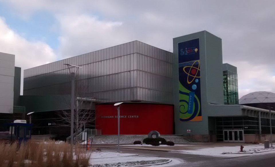 The Michigan Science Center building with its red entrance wall, colourful banner and the dome at the right edge on a snowy day