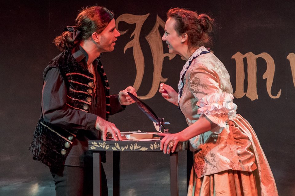 An actor and actress in period costume lean towards each other over a table with a quill pen on stage