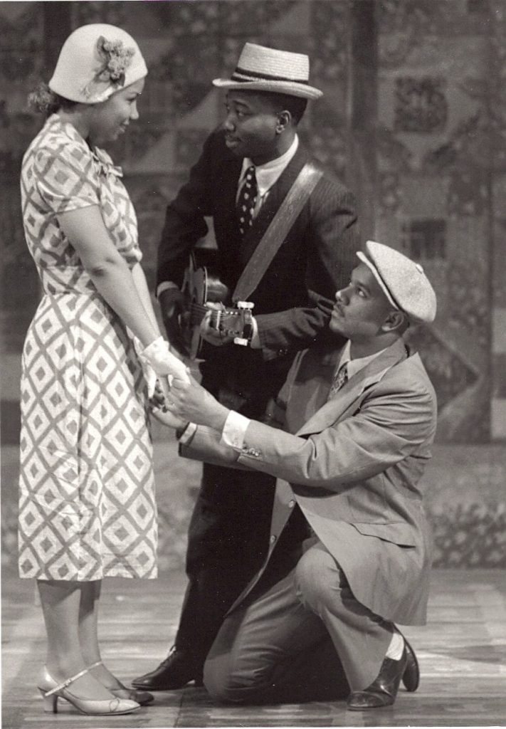Black-and-white production photo of a man in a cap kneeling before a woman in a patterned dress, with a guitarist behind them